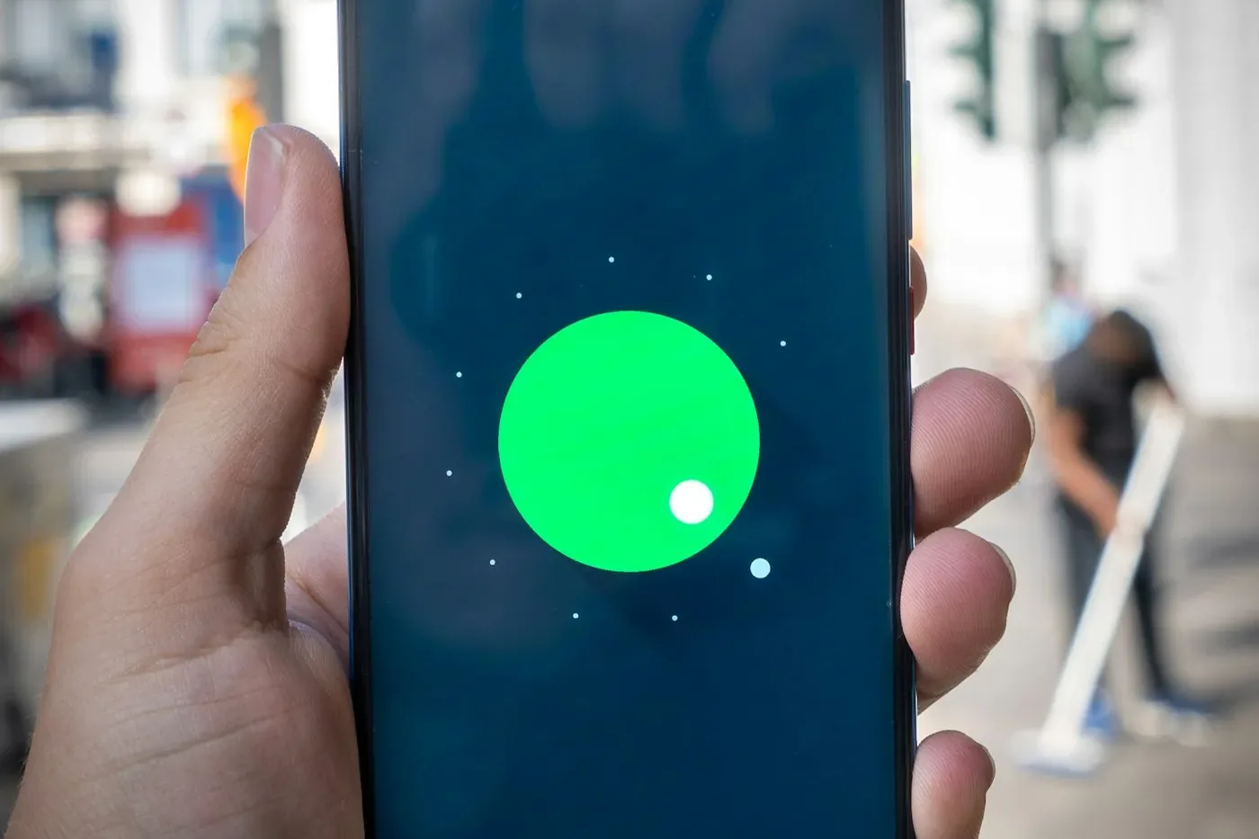 Hand hält ein Smartphone, auf dem dunklen Display ein großer grüner Kreis, im Hintergrund eine unscharfe Straßenszene